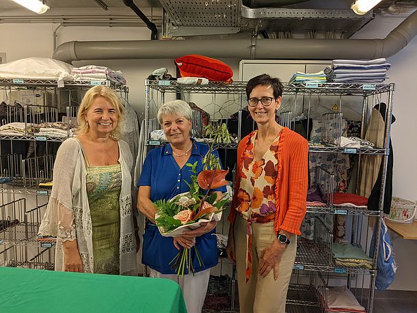 Drei Damen vor den Wäschewägen. Die Damen in der Mitte hält einen Blumenstrauß.